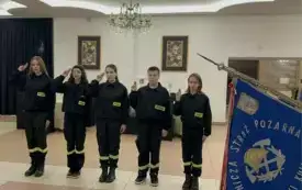 Na pierwszym planie niebieska płachta sztandaru OSP Złotniki Kujawskie. W tle pięciu młodych druch&oacute;w Młodzieżowej OSP salutuje