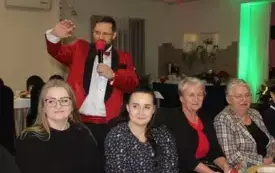 Cztery kobiety siedzą przy wigilijnym stole. Za nimi Krzysztof Filasiński z mikrofonem w ręku śpiewa kolędę. Drugą ręką macha w geście pozdrowienia.