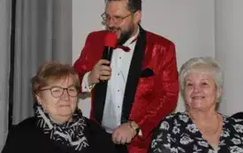 Dwie kobiety siedzą przy wigilijnym stole. Za nimi tenor Krzysztof Filasiński z mikrofonem w ręku śpiewa kolędę.