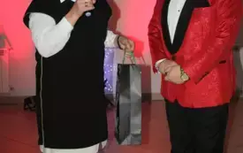 Kobieta i mężczyzna elegancko ubrani stoją na środku restauracyjnej sali. Kobieta z mikrofonem w ręku. W drugiej trzyma upominek dla mężczyzny zapakowany w świąteczną paczkę.