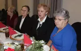 Cztery kobiety ubrane odświętnie siedzą z poważnymi minami za stołem. Na stole biały obrus i zastawa z dekorcją ze świerkowej gałązki na talerzach.