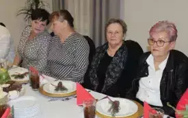Cztery kobiety siedzą przy stole. Dwie z nich rozmawiają. Na stole zastawa i biały obrus. Na talerzykach dekoracja z gałąski świekowej iczerwonej kokardy.