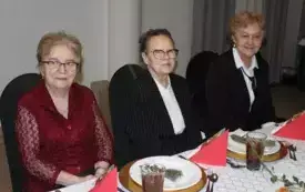 Trzy kobiety elegansko, odświetnie ubrane siedzą przy stole nakrytym białym obrusem. Na stole zastawa i świąteczna dekoracja z gałązek świeku.