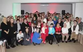 Bardzo duża grupa kobiet i kulku mężczyzn pozuje na środku restauracyj ej sali do zdjęcia. Część z nich stoi, część siedzi na krzesłach a część kuca. Wszyscy odświętnie, elegancko ubrani. Uśmiechają się.