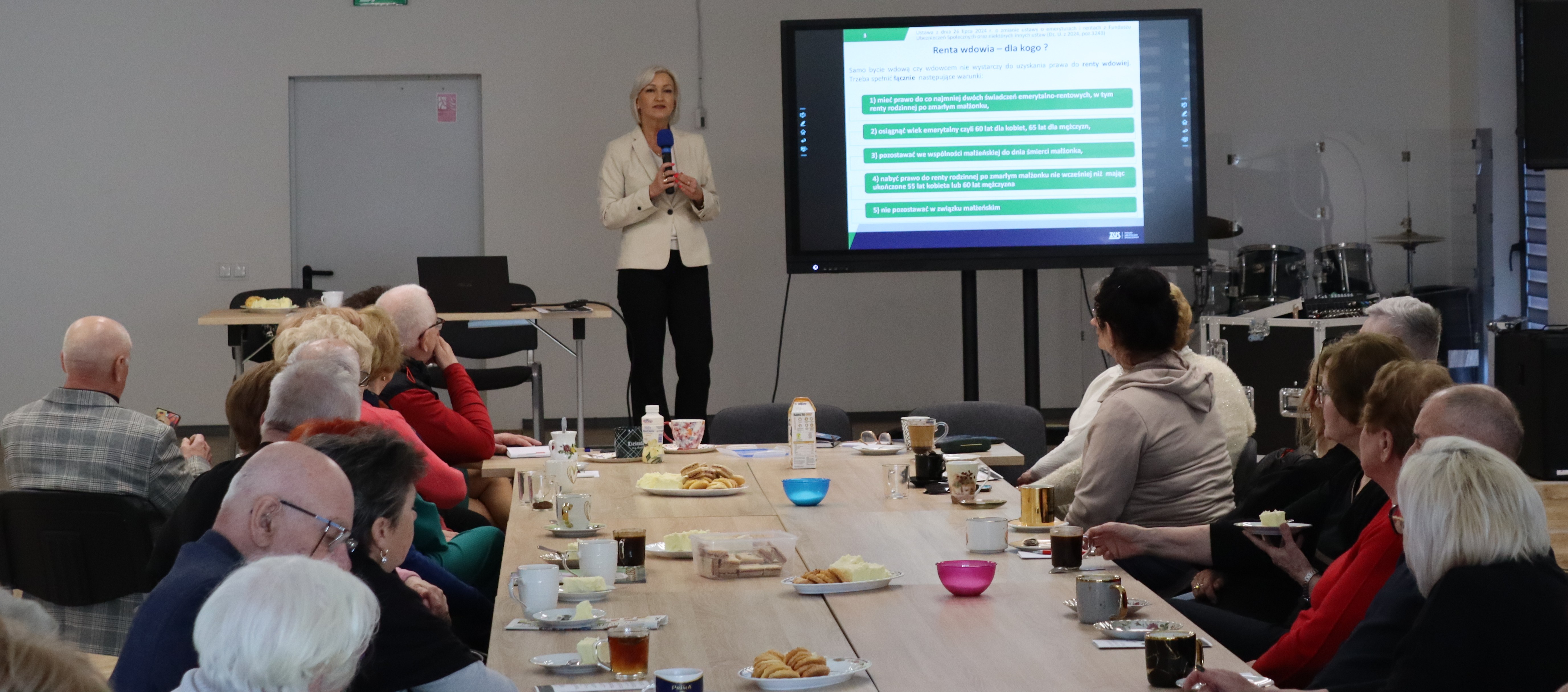 Kobieta z&nbsp;mikrofonem stoi i&nbsp;przemawia do siedzących przy stole os&oacute;b